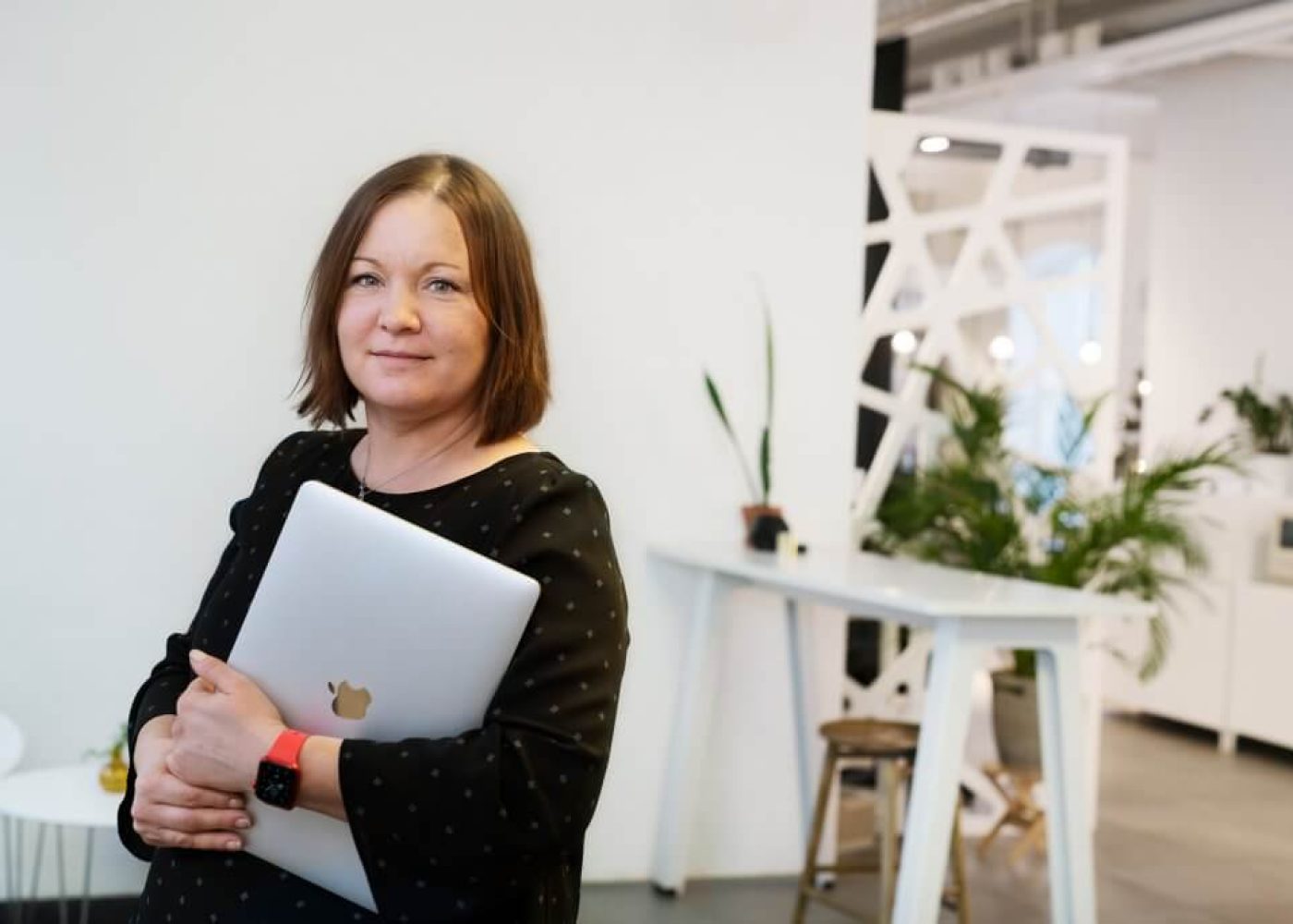 En kvinna med axellångt brunt hår, klädd i svart topp och röd smartklocka, står inomhus och håller i en stängd silverfärgad laptop. Moderna kontorsmöbler och växter syns i bakgrunden.