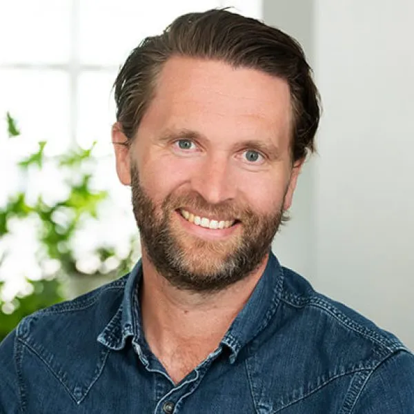 En leende man med brunt hår och skägg, klädd i en blå jeansskjorta, sitter inomhus med en suddig bakgrund med en växt och ett fönster.