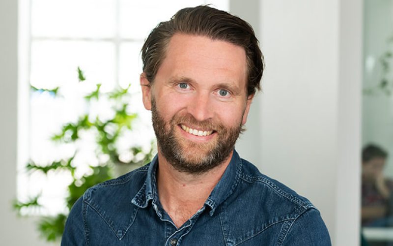 En leende man med brunt hår och skägg, klädd i en blå jeansskjorta, sitter inomhus med en suddig bakgrund med en växt och ett fönster.