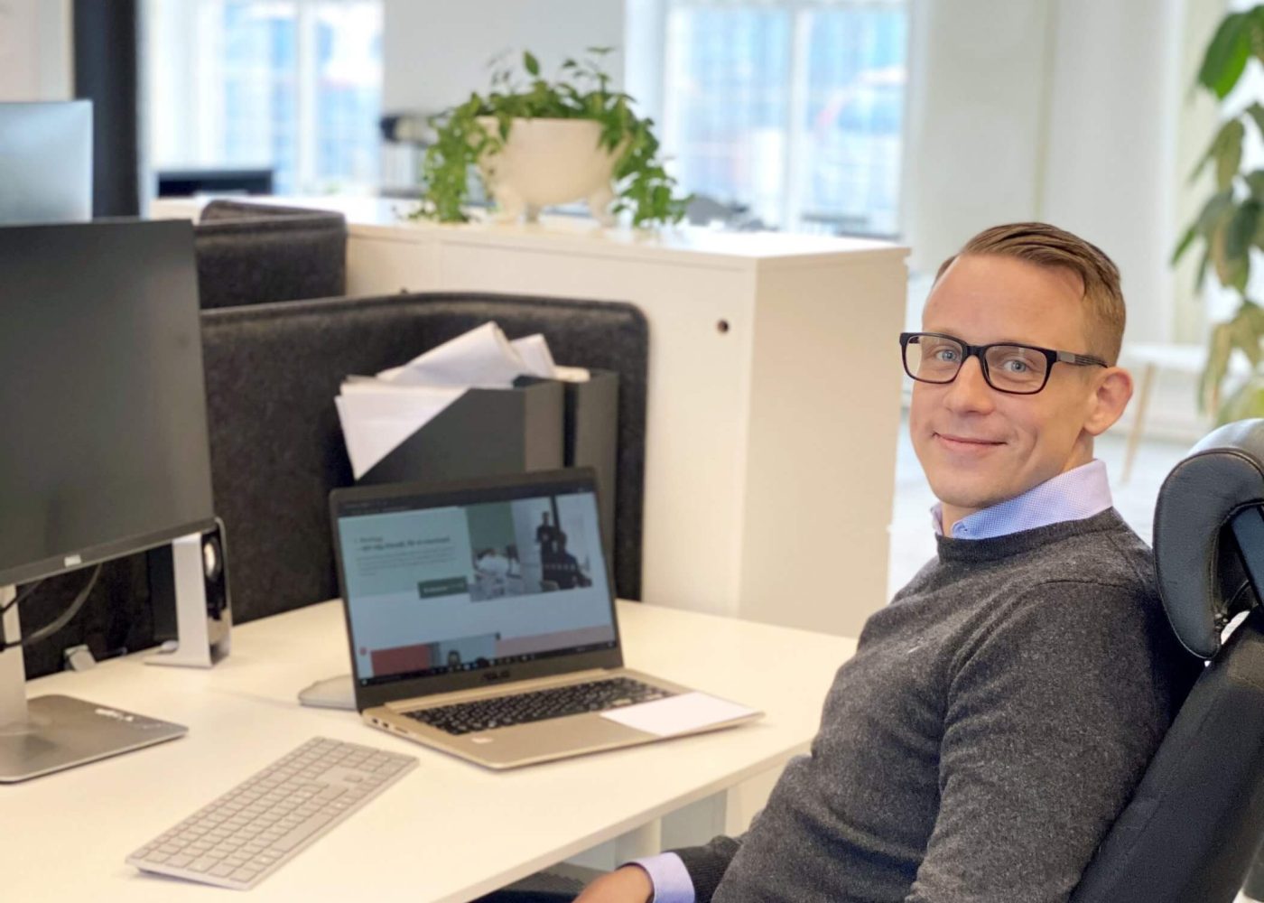 En man med glasögon och grå tröja sitter vid ett skrivbord på ett modernt kontor och ler mot kameran. En bärbar dator, ett tangentbord och en bildskärm står på skrivbordet, med stora fönster och växter i bakgrunden.