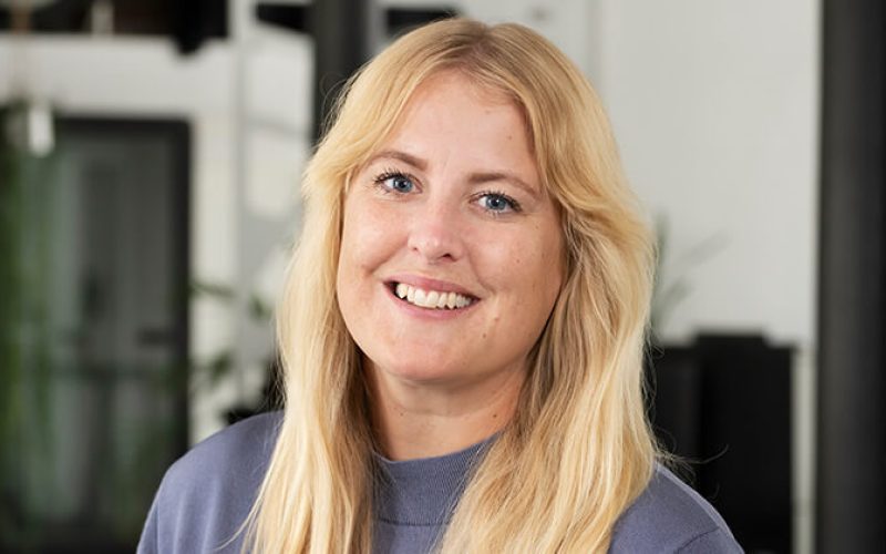 Leende kvinna med långt blont hår och grå topp står inomhus mot en suddig bakgrund med fönster och växter.