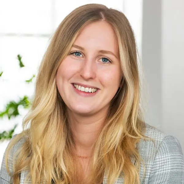 En ung kvinna med långt blont hår och en grårutig kavaj ler mot kameran. Hon befinner sig inomhus med ett ljust fönster och några gröna växter i bakgrunden.