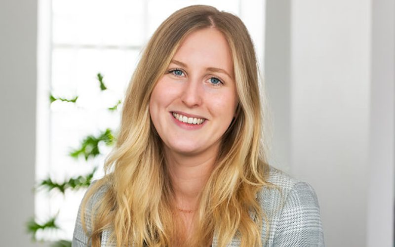 En ung kvinna med långt blont hår och en grårutig kavaj ler mot kameran. Hon befinner sig inomhus med ett ljust fönster och några gröna växter i bakgrunden.