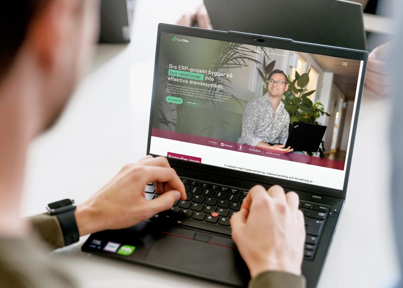 En person använder en bärbar dator och surfar på en webbplats med en leende man som håller i en bärbar dator och text om att bygga effektiva affärssystem. Andra personer och bärbara datorer syns i bakgrunden.