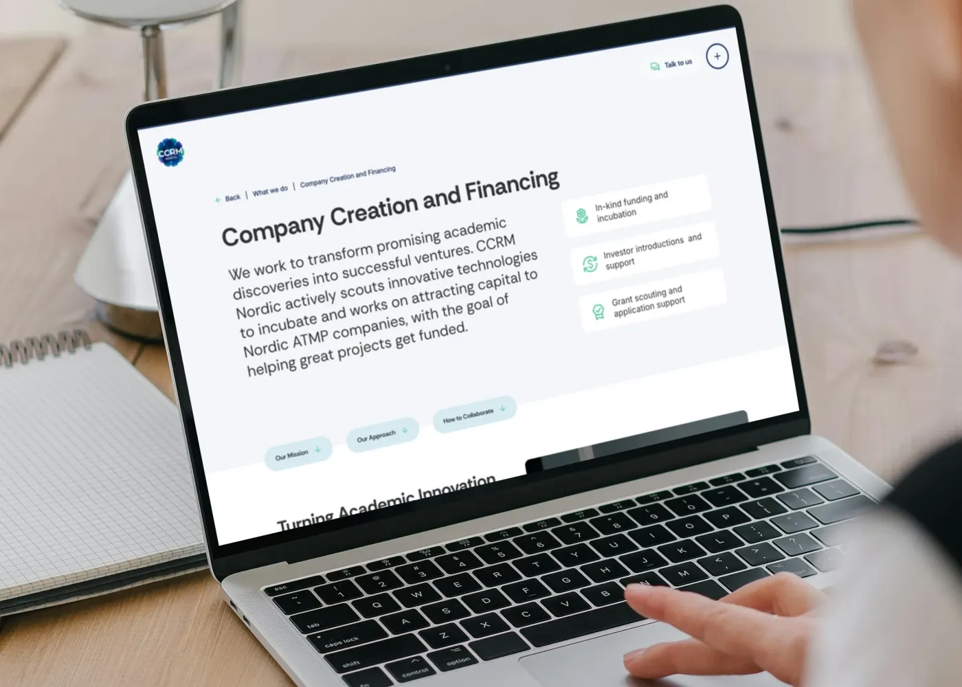 En person använder en bärbar dator som visar en webbsida om företagsbildning och finansiering, med information om akademisk innovation, finansiering och projektstöd. Den bärbara datorn står på ett skrivbord av trä med en lampa i bakgrunden.
