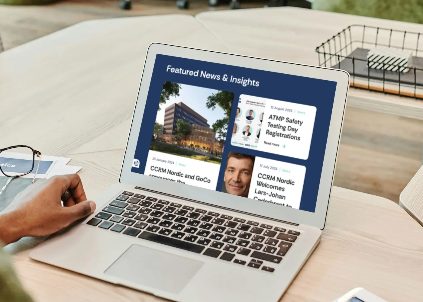 En person använder en bärbar dator som visar en webbsida med rubriken Featured News & Insights och flera nyhetsartiklar, placerad på ett träbord med papper och en trådkorg i närheten.
