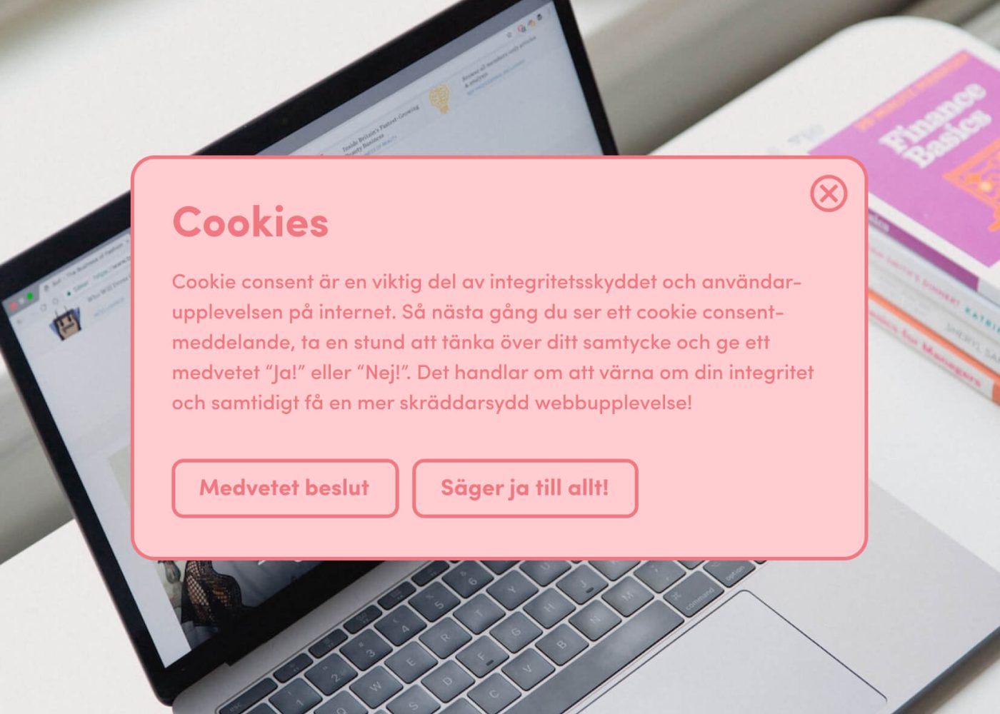 En rosa cookie consent popup på svenska visas på en laptopskärm, med knappar som säger Medvetet beslut och Säger ja till allt! Bredvid den bärbara datorn ligger böcker, bland annat en med lila omslag.