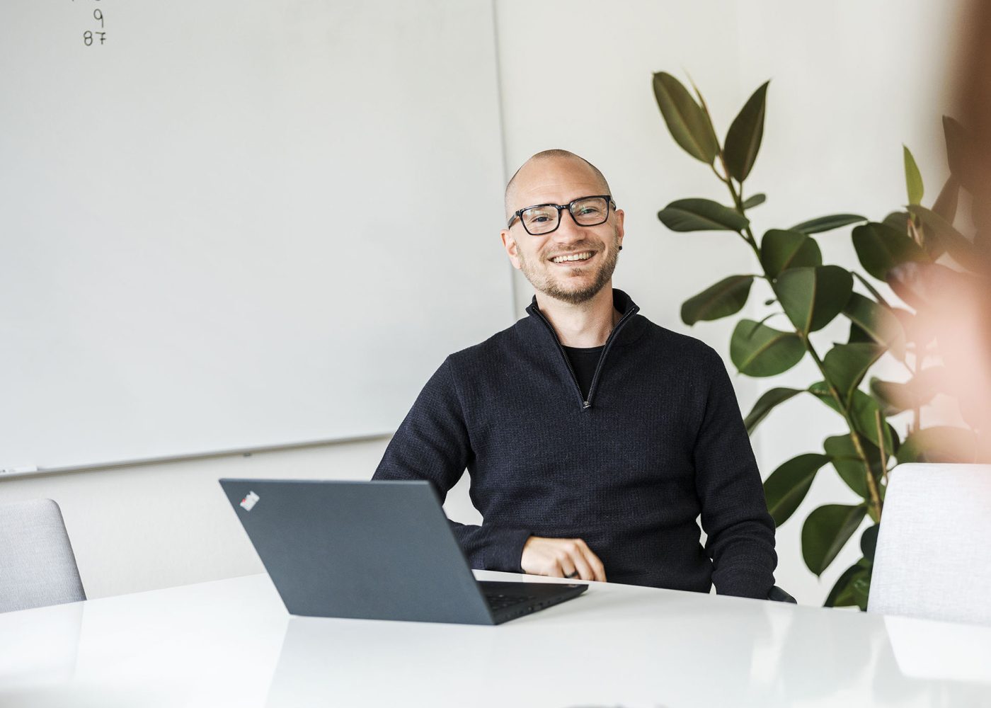 En leende person med glasögon sitter vid ett vitt bord med en öppen laptop, i ett ljust kontor med en bladväxt och en whiteboard med siffror skrivna på i bakgrunden.