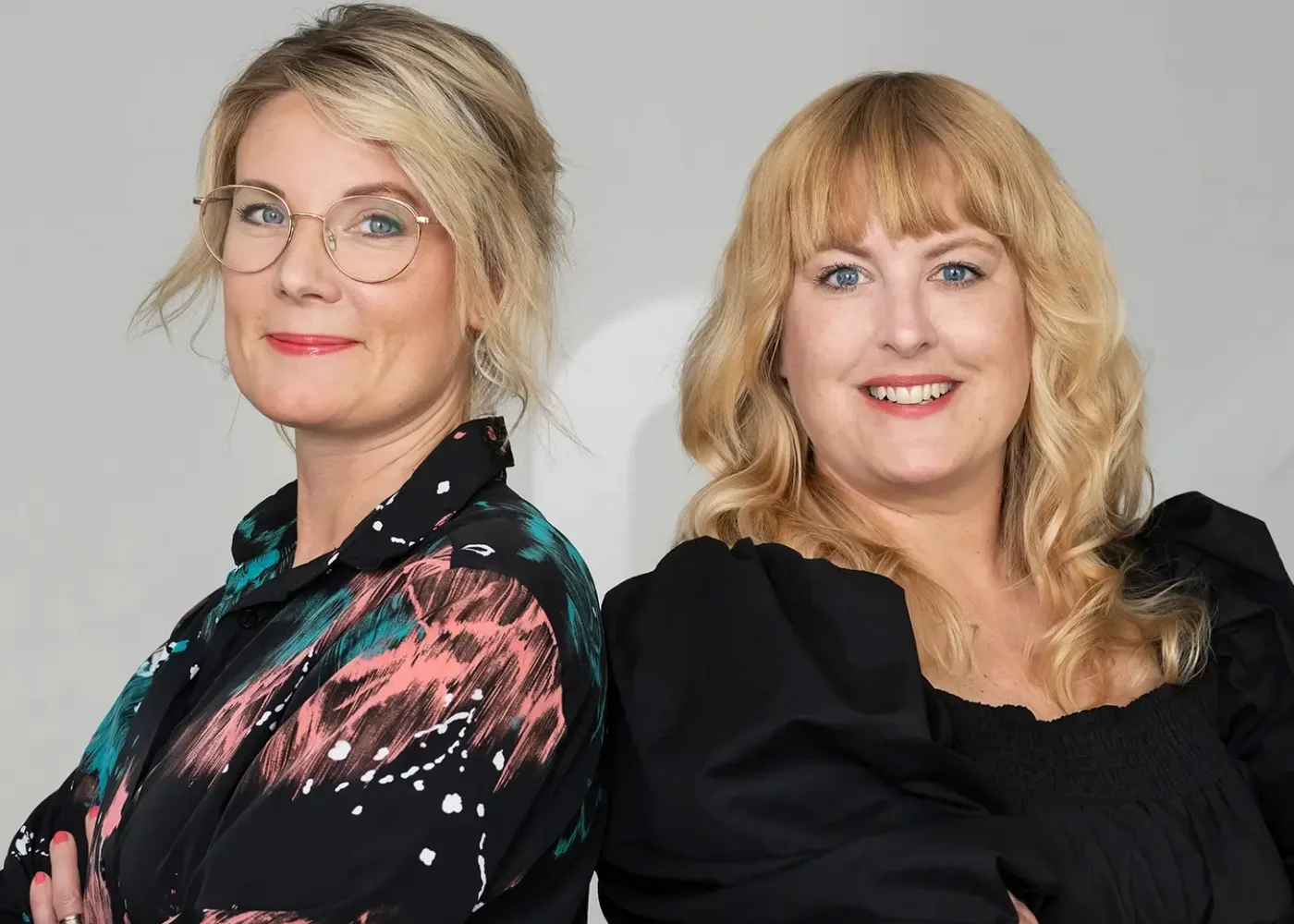 Två kvinnor står rygg mot rygg och ler mot kameran. Den ena har blont hår i lösa vågor och bär en svart klänning, medan den andra har ljust hår i en knut, glasögon och en färgstark mönstrad blus.