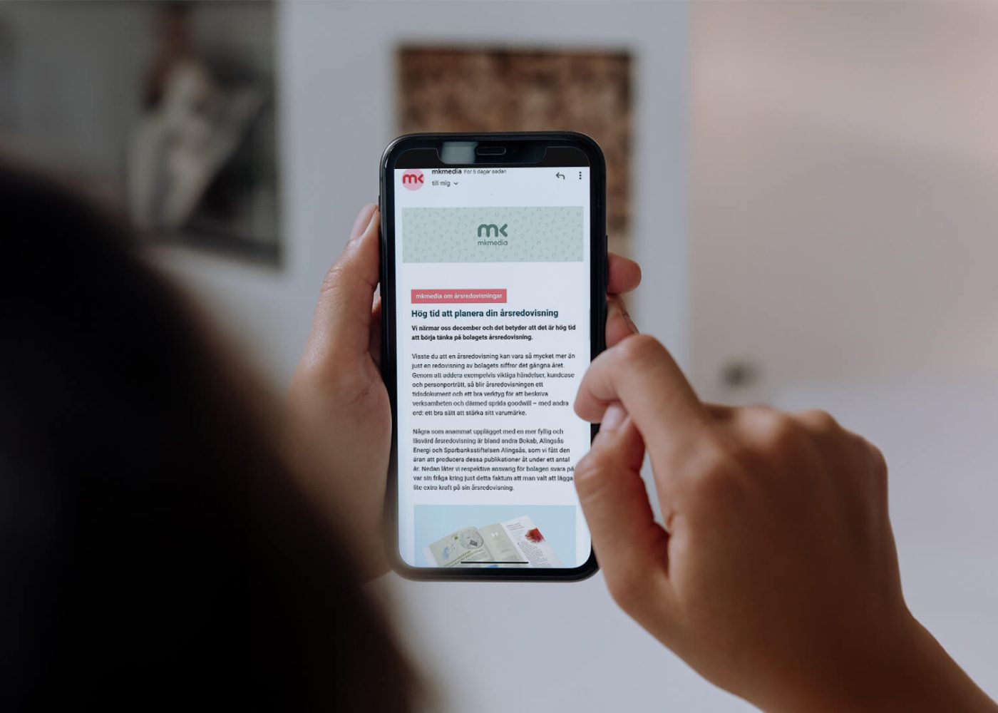 En person håller i en smartphone och bläddrar igenom en artikel eller ett meddelande med text och en liten bild på skärmen, inomhus med suddig bakgrund.