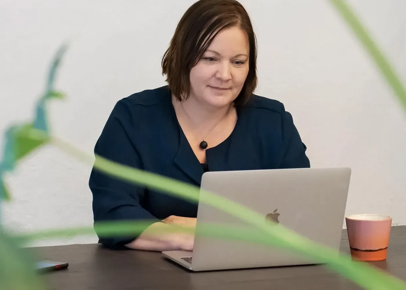 En kvinna med kort brunt hår arbetar på en laptop vid ett mörkt bord, med en rosa kaffekopp i närheten. Gröna växtblad syns i förgrunden, något oskarpa. Bakgrunden är enfärgad och ljus.