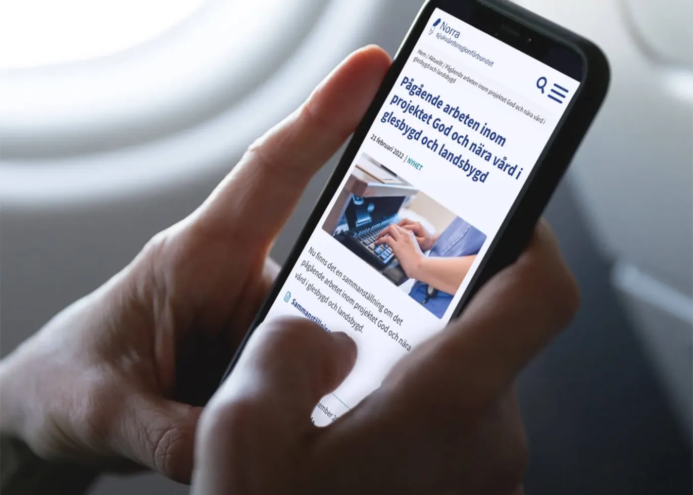 En person sitter vid ett flygplansfönster med en smartphone i handen och surfar på en svensk webbplats om pågående arbete i vårdprojekt; på skärmen syns text och en bild av händer som använder en surfplatta.