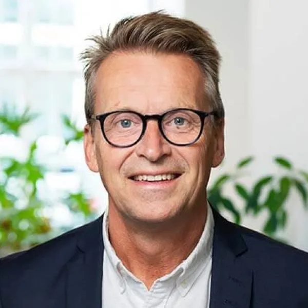 En leende medelålders man med glasögon och kort, ljusbrunt hår, klädd i mörk kavaj och vit skjorta, står inomhus med gröna växter och stora fönster i bakgrunden.