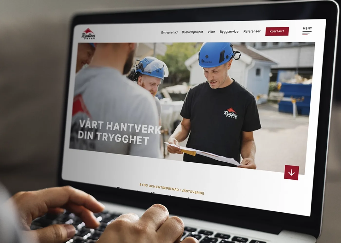 En person skriver på en bärbar dator som visar en webbplats med en bild av byggnadsarbetare som granskar papper utomhus. Webbplatsen är på svenska och har texten Vårt hantverk, din trygghet.