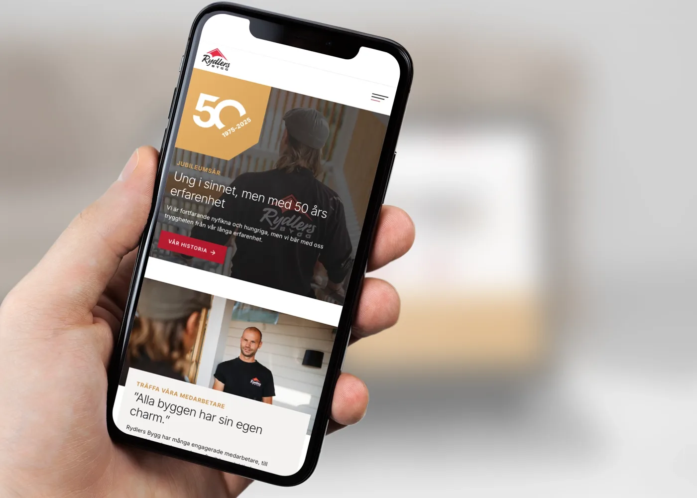 En hand håller i en smartphone som visar en webbplats med logotypen Rydlers, en 50-årsbanner och bilder på människor. Webbplatsen förefaller innehålla artiklar på svenska om erfarenhet och individualitet i byggandet.