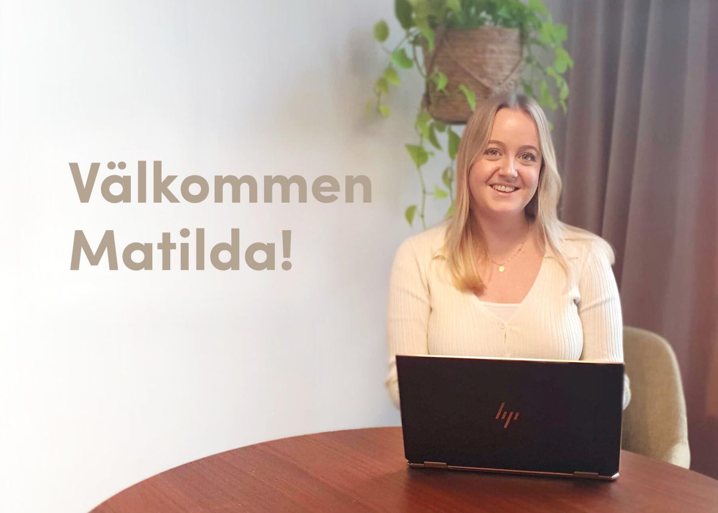 En leende kvinna sitter vid ett runt bord med en bärbar dator av märket HP. Bakom henne hänger en växt på väggen. Texten på bilden säger Välkommen Matilda! i stora, ljusa typsnitt.
