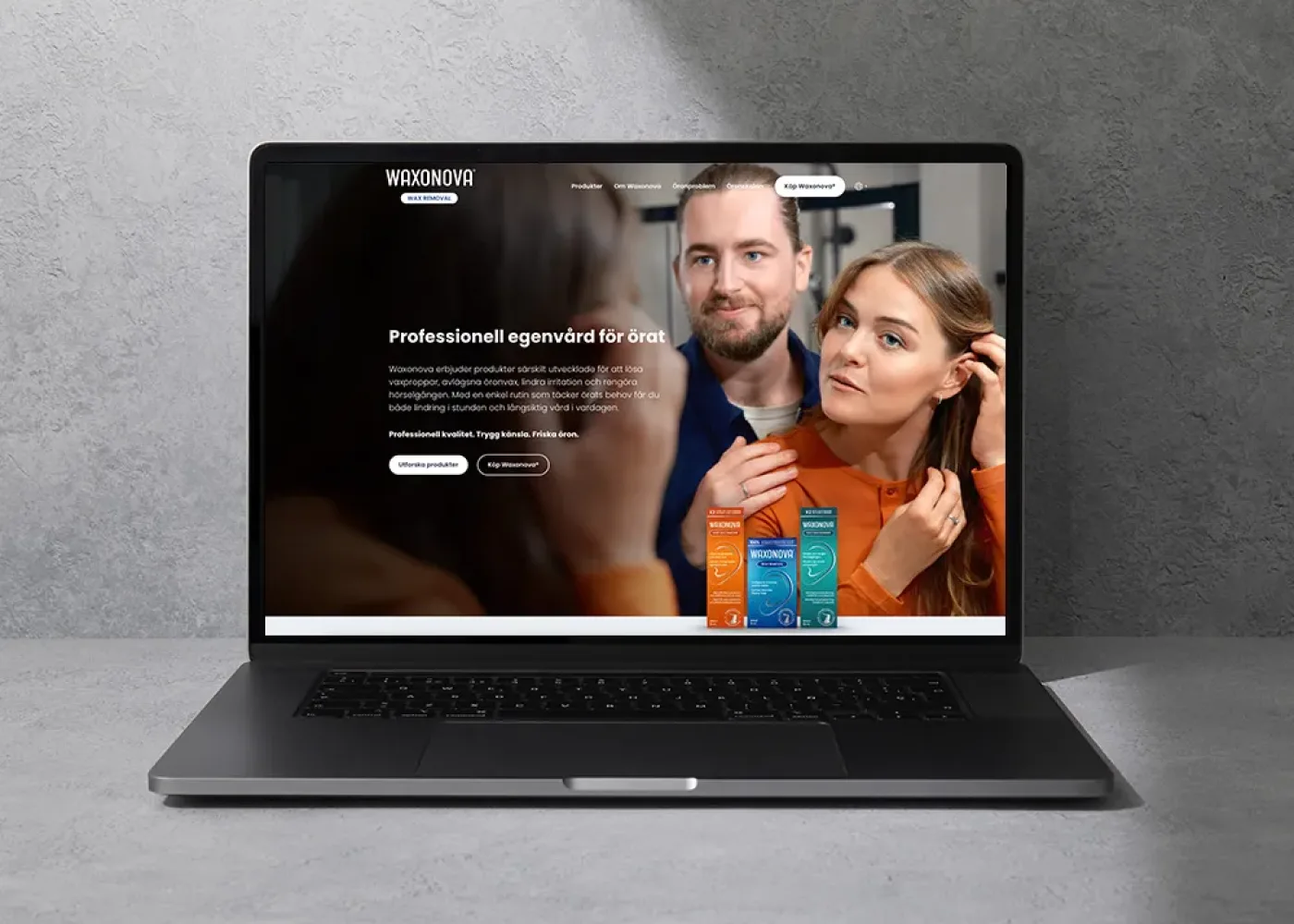 En svart bärbar dator på en grå yta visar en webbplats med en man som hjälper en kvinna att ta på sig en orange jacka och flera färgglada produktlådor visas längst ner på skärmen.
