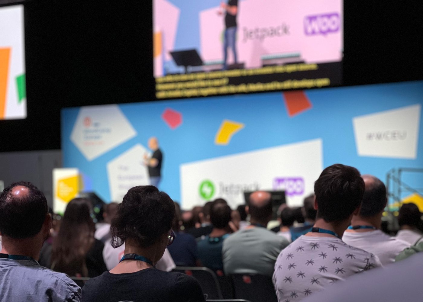 Åhörare ser en talare presentera på scenen på en konferens, med en ljusblå bakgrund med logotyper som Jetpack och WOO, och hashtaggen #WCEU synlig till höger.