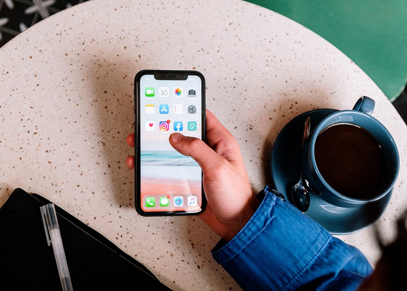 En person som håller i en smartphone med synliga appikoner, sittande vid ett runt bord med en kopp kaffe och en svart anteckningsbok samt en penna.